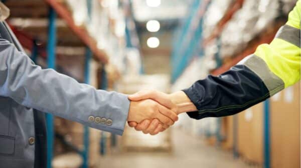 Händedruck mit Arm einer Person in gelb-schwarzer Logistikjacke und mit Arm einer Person in grauem Anzug, Hintergrund hohe Regale unscharf