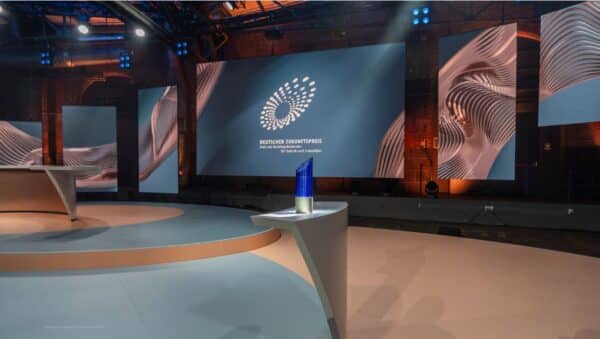 leeres TV-Studio mit rundem Tisch, auf dem die blaue Trophäe des Deutschen Zukunftspreises steht; an der Wand im Hintergrund weißes Logo des Preises