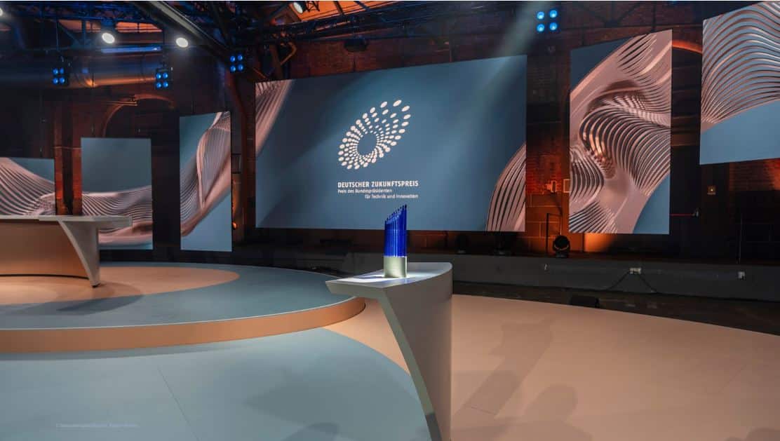 leeres TV-Studio mit rundem Tisch, auf dem die blaue Trophäe des Deutschen Zukunftspreises steht; an der Wand im Hintergrund weißes Logo des Preises